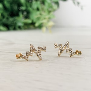 Puede incluir: Un par de pendientes de oro con un corazón y un diseño de EKG de diamantes de imitación.