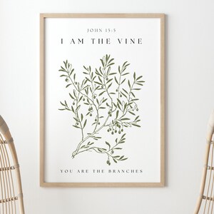 A framed print with a white background and a green illustration of a vine with leaves and berries. The text on the print reads 'John 15:5', 'I AM THE VINE', and 'YOU ARE THE BRANCHES'. The print is hanging on a white wall between two hanging wicker chairs. The chairs are hanging from the ceiling and are made of natural woven material. The chairs are empty and facing away from each other. There is a vase with dried grass in front of the chairs.
