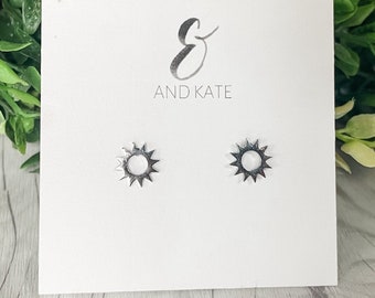 Peut inclure: Une paire de boucles d'oreilles soleil en argent avec des pierres d'opale blanche. Les boucles d'oreilles sont sur une carte blanche avec le texte "& AND KATE" imprimé dessus.