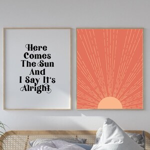 Könnte beinhalten: Zwei gerahmte Drucke an einer weißen Wand über einem Bett mit einer weißen Bettdecke und einem Kopfteil aus Holz. Der Druck links sagt "Here Comes The Sun And I Say It's Alright". Der Druck rechts ist eine stilisierte orange Sonne mit Strahlen.