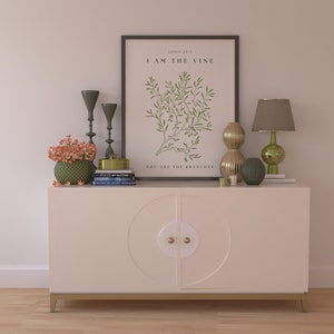 A white cabinet with gold accents and two doors. The cabinet is decorated with a circular design on each door. The cabinet is sitting on a wooden floor. On top of the cabinet are a variety of decorative items, including a framed print with the text 'John 15:5 I AM THE VINE YOU ARE THE BRANCHES', two green candlesticks, a gold box, a stack of books, a green vase with orange flowers, a gold and green lamp, and a gold and green vase.