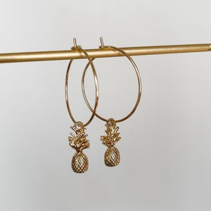 Könnte beinhalten: Ein Paar goldene Creolen mit Ananas-Anhängern. Die Ohrringe hängen an einem goldenen Ständer.