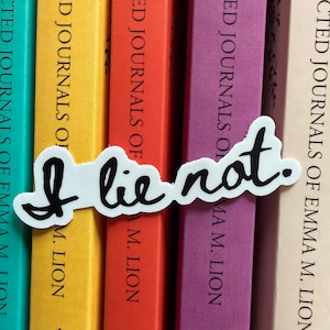 May include: A white die-cut sticker featuring the black handwritten text I lie not. The sticker is positioned horizontally across the spines of five books with covers in shades of teal, yellow, orange, purple, and light beige, all titled Collected Journals of Emma M. Lion.