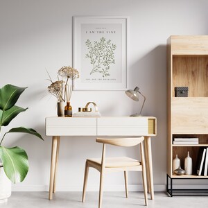 A white desk with two drawers and wooden legs. A wooden chair with a light brown cushion is in front of the desk. A framed print with the text 'John 15:5 I AM THE VINE YOU ARE THE BRANCHES' is hanging on the wall behind the desk. A lamp with a gray shade is on the desk. A small wooden shelf with books and decorative items is on the right side of the image.