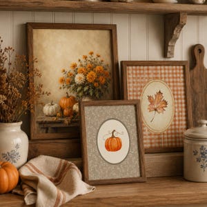 May include: Three framed wall art pieces featuring autumn themes. One print shows a bouquet of orange flowers and pumpkins on a table. Another displays a single maple leaf inside an oval on a gingham background. The third shows a pumpkin illustration.