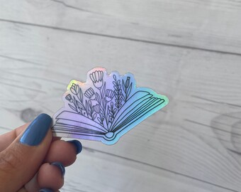 May include: A holographic sticker of an open book with flowers growing out of it. The sticker is outlined in black and has a rainbow iridescent finish.