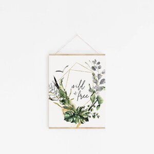 May include: A white poster with a gold geometric frame and green and grey watercolour leaves. The text "wild & free" is written in black inside the frame.