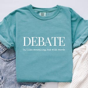 May include: A teal Comfort Colors t-shirt with the word "DEBATE" in white, and the definition: "(n.) like wrestling, but with words." The shirt is a solid colour and has a crew neck.