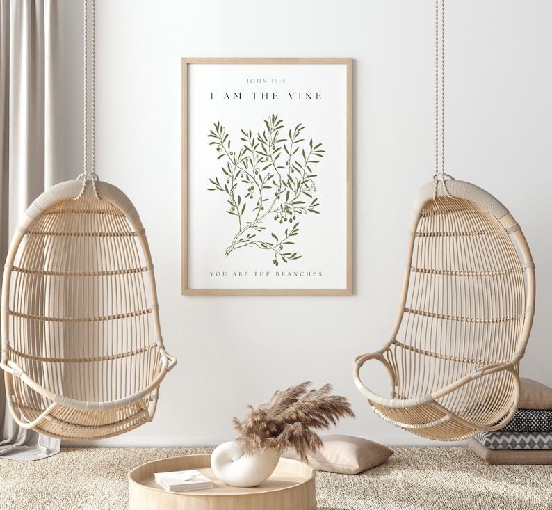 A framed print with a white background and a green illustration of a vine with leaves and berries. The text on the print reads 'John 15:5', 'I AM THE VINE', and 'YOU ARE THE BRANCHES'. The print is hanging on a white wall between two hanging wicker chairs. The chairs are hanging from the ceiling and are made of natural woven material. The chairs are empty and facing away from each other. There is a vase with dried grass in front of the chairs.