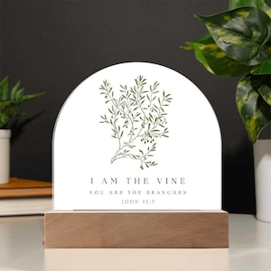 May include: A white arched plaque with a green olive branch illustration and the text "I AM THE VINE YOU ARE THE BRANCHES JOHN 15:5". The plaque is mounted on a rectangular wooden base. The background includes a plant.