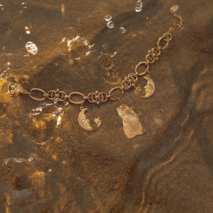 May include: A gold-colored bracelet with moon and star charms, resting on a sandy surface with water droplets. The bracelet features a delicate chain design and celestial-themed pendants.