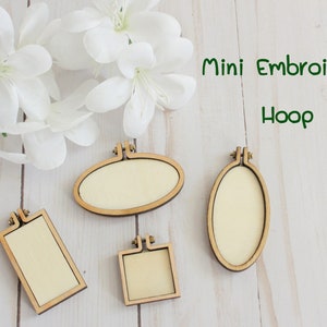 Peut inclure: Quatre cerceaux à broder en bois de différentes tailles et formes. Deux sont ovales, un est carré et un est rectangulaire. Les cerceaux sont sur une surface en bois blanc. Le texte "Mini Embroidery Hoop" est écrit en vert au-dessus des cerceaux.