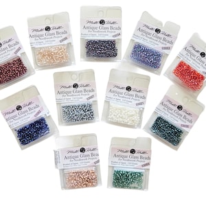 Può includere: Una collezione di piccole perline di vetro antiche colorate in varie tonalità, tra cui rosso, blu e bianco. Ogni confezione è etichettata "Antique Glass Beads" ed è progettata per progetti di ricamo. Le perline sono confezionate in piccoli sacchetti di plastica trasparente.