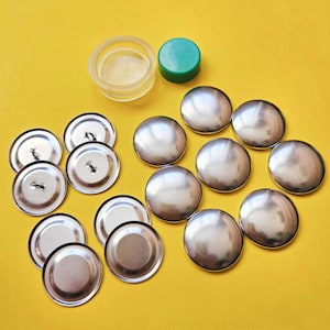 May include: A set of silver-colored metal buttons with a green plastic cap and a clear plastic container on a yellow background.