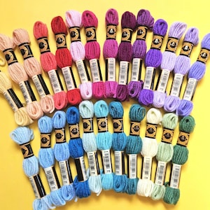 May include: A selection of embroidery floss skeins in a range of colours, including pinks, purples, blues, greens, and creams, arranged on a yellow surface. Each skein has a black and white label.