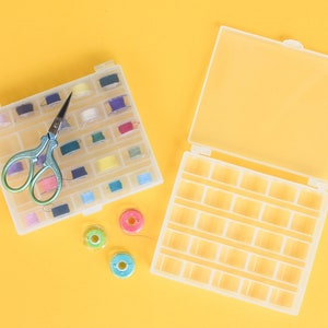 May include: Two clear plastic storage boxes with compartments for small items, such as thread spools. The boxes are open and empty, except for one box that has colourful thread spools and a pair of small scissors.