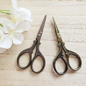 Könnte beinhalten: Zwei Paare verzierter Scheren im antiken Stil mit dekorativen Griffen. Die Scheren liegen auf einer hellbraunen Holzoberfläche.