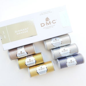 May include: A collection of DMC Diamant Grande metallic embroidery threads in various colors, including gold, silver, and bronze. The threads are displayed with their packaging, which features the DMC logo and the words "Diamant Grande."