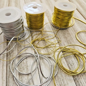 May include: Two spools of metallic cord, one gold and one silver, are unwound on a wooden surface. The cord is braided and has a subtle texture.