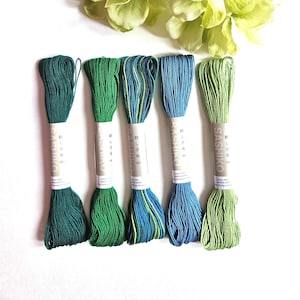May include: Five skeins of Sashiko embroidery thread in shades of green and blue, with Japanese text on the labels. The threads are arranged below a cluster of light green hydrangea flowers, all on a white background.