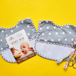 Puede incluir: Dos baberos grises con lunares blancos y ribete de cuadros, junto a un paquete etiquetado como "Baby Bib" e hilo de bordar. Los baberos tienen cierre de velcro. El paquete muestra a un bebé siendo alimentado.