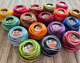May include: A collection of 15 colorful embroidery floss spools with white and colored stripes. The spools are arranged in a circle on a wooden surface. The spools are labeled with the text "DMC Coton Perle 8/5 100m".