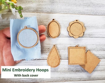 May include: Six wooden embroidery hoops of various shapes, including round, oval, teardrop, and square, with a light blue fabric in one of the round hoops. The hoops are on a white wooden surface. The text "Mini Embroidery Hoops With back cover" is at the bottom of the image.