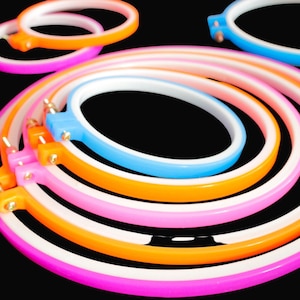 May include: A set of five embroidery hoops in various sizes. The hoops are made of plastic and have a white inner ring and a coloured outer ring. The colours are pink, orange, light blue, and a darker pink.