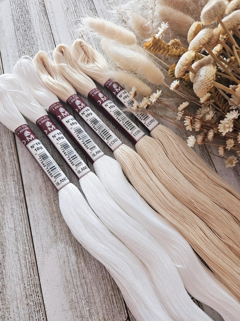 May include: A collection of embroidery floss in shades of white and beige, arranged on a wooden surface. Each spool is labeled with the brand name, thread weight, and color. Dried flowers and foliage are in the background.
