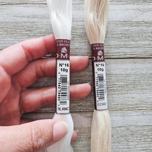 May include: Two spools of DMC cotton embroidery floss, one white and one ecru, are held against a wooden background. Each spool is labeled with the brand, weight (10g), and color name. The white spool is labeled "BLANC". The ecru spool is labeled "ECRU".