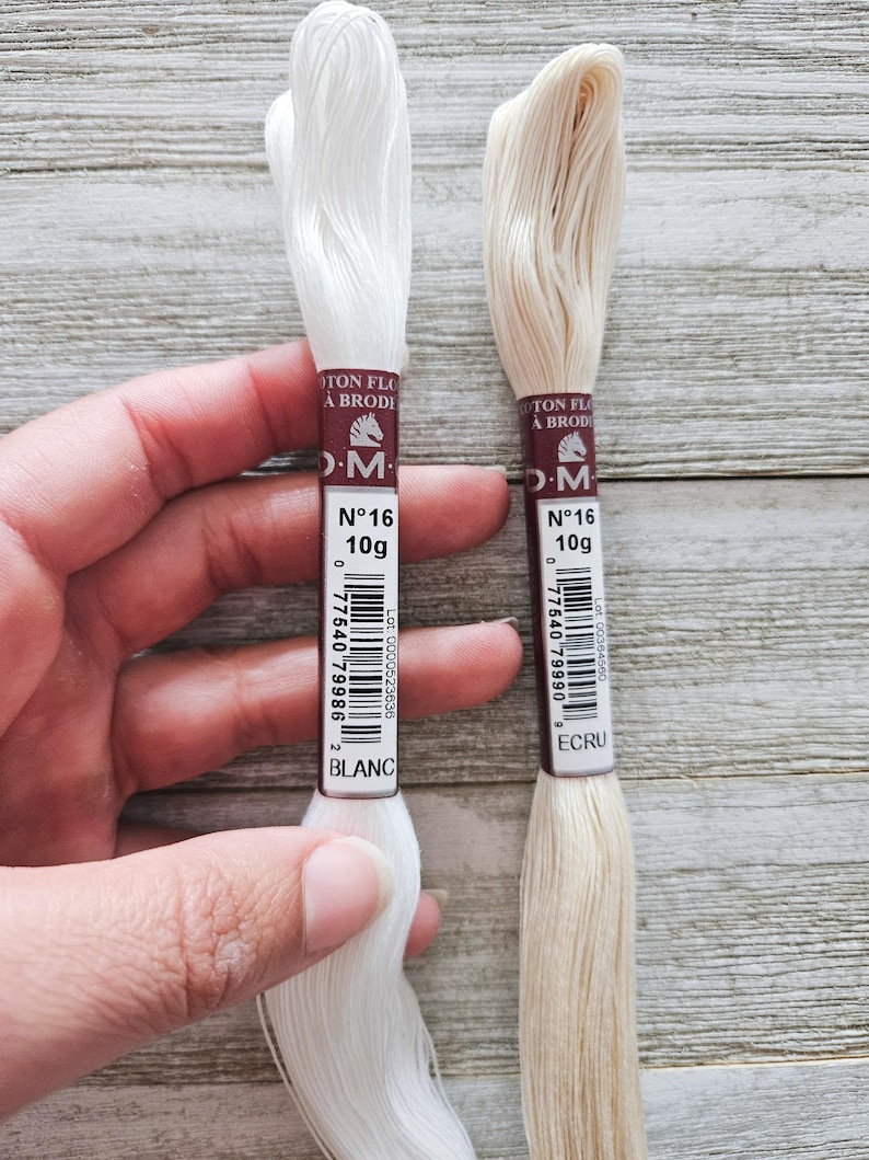 May include: Two spools of DMC cotton embroidery floss, one white and one ecru, are held against a wooden background. Each spool is labeled with the brand, weight (10g), and color name. The white spool is labeled "BLANC". The ecru spool is labeled "ECRU".
