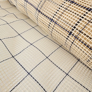May include: A rolled-up rug with a grid pattern. The rug is made of a beige woven material with a dark blue grid design. The grid is made of thick lines that intersect to form squares.