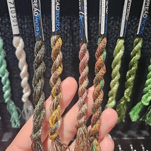 May include: A selection of Threadworx artisan-dyed embroidery floss, featuring a range of colours with metallic threads. The image shows the floss held in a hand, highlighting the braided texture. Labels read "Kreinik Fine #8 Braid Artisan Dyed".