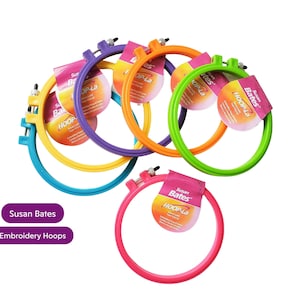 May include: A set of five colourful plastic embroidery hoops with the brand name "Susan Bates Hoop-La" printed on the packaging. The hoops are in various colours, including yellow, green, blue, purple, and pink.