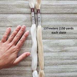 May include: Two skeins of embroidery floss, one white and one ecru, are displayed against a wooden background. Each skein is labeled with the brand, color, and weight. The text "137 meters (150 yards) each skein" is also visible.