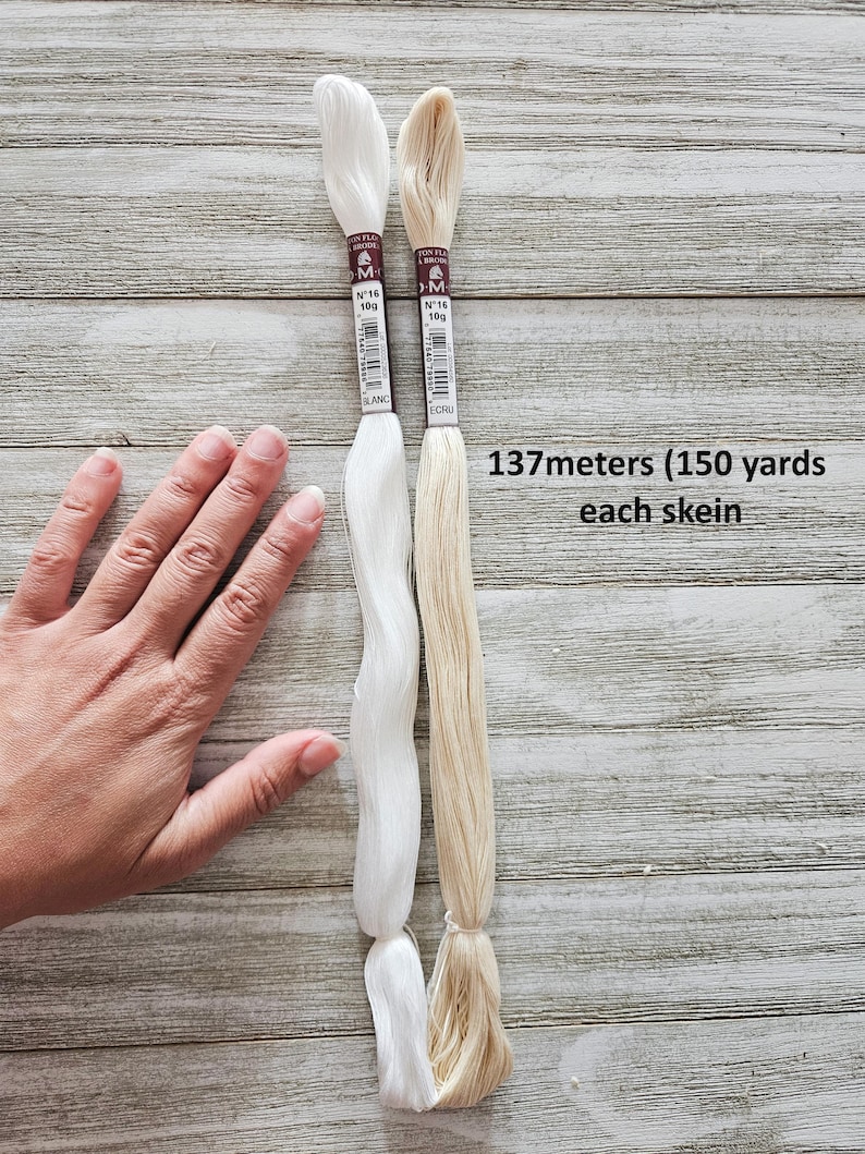 May include: Two skeins of embroidery floss, one white and one ecru, are displayed against a wooden background. Each skein is labeled with the brand, color, and weight. The text "137 meters (150 yards) each skein" is also visible.