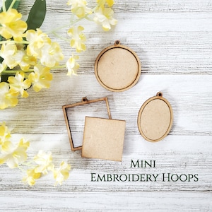 May include: Three wooden embroidery hoops of different shapes and sizes. The hoops are on a white wooden surface. The text "MINI EMBROIDERY HOOPS" is printed below the hoops.