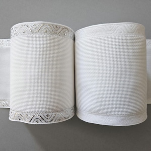 Puede incluir: Dos rollos de cinta de tela blanca con un patrón de rejilla texturizado en el centro y bordes decorativos. El rollo izquierdo muestra un diseño ondulado de color plata metálica con pequeños acentos circulares, mientras que el derecho tiene un sutil patrón de ondas en relieve.