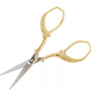 May include: Gold-colored embroidery scissors with ornate handles and silver blades. The scissors have a decorative floral design on the handles and are open, ready for use. The scissors are designed for sewing and crafting.