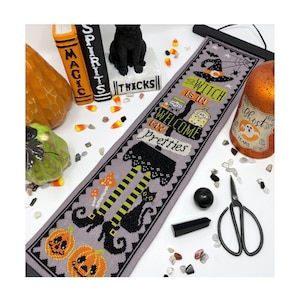 May include: A cross-stitch banner with a Halloween theme, featuring the text "The Witch is in, Welcome My Pretties." The design includes a witch's hat, legs, pumpkins, and a black cat. The banner is displayed with Halloween decorations.