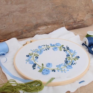 May include: A circular wooden embroidery hoop containing white fabric with a hand-stitched wreath of blue ribbon roses, small dark blue buds, and green embroidered leaves. A spool of blue ribbon and a bundle of green embroidery thread are nearby.