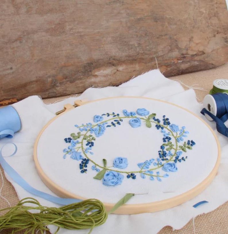 May include: A circular wooden embroidery hoop containing white fabric with a hand-stitched wreath of blue ribbon roses, small dark blue buds, and green embroidered leaves. A spool of blue ribbon and a bundle of green embroidery thread are nearby.