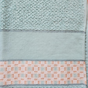 May include: A close-up of a light teal bath towel. The towel has a textured surface with a woven border featuring a geometric pattern in shades of peach, white, and teal. The towel is made of soft material.