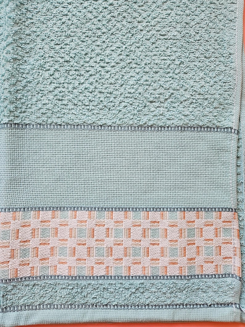 May include: A close-up of a light teal bath towel. The towel has a textured surface with a woven border featuring a geometric pattern in shades of peach, white, and teal. The towel is made of soft material.