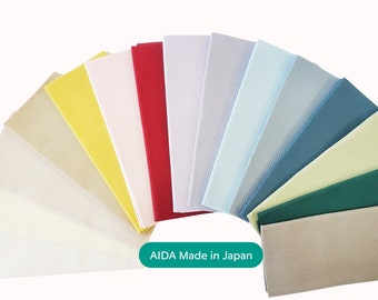 Può includere: Campioni di tessuto AIDA in una varietà di colori, tra cui beige, bianco, giallo, rosso, blu chiaro, blu scuro, verde e marrone. Il tessuto è disposto a ventaglio con il testo "AIDA Made in Japan" in basso.