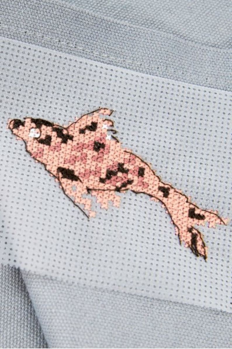 May include: A pink and brown embroidered fish design on a light blue fabric background.