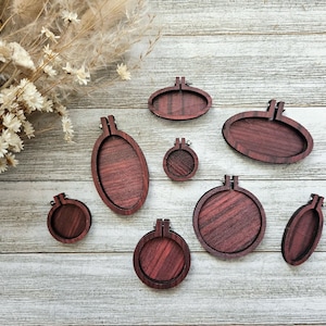 May include: A selection of unfinished wooden embroidery hoops in various sizes and shapes, in reddish-brown hues. They are displayed on a light grey wooden surface, with dried flowers in the top left corner.