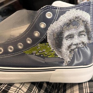 May include: Blue canvas high-top sneakers with a white sole and a black trim. The sneakers have a mesh design and feature a cross-stitch image of a person's face.