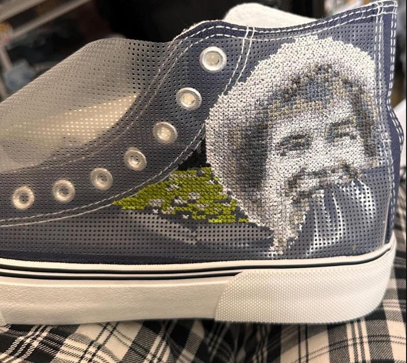 May include: Blue canvas high-top sneakers with a white sole and a black trim. The sneakers have a mesh design and feature a cross-stitch image of a person's face.