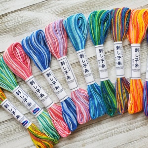 May include: A collection of nine skeins of variegated Sashiko embroidery thread, each featuring a white paper label with Japanese text. The threads display a spectrum of colours including green, pink, blue, orange, and purple, with multi-coloured strands.